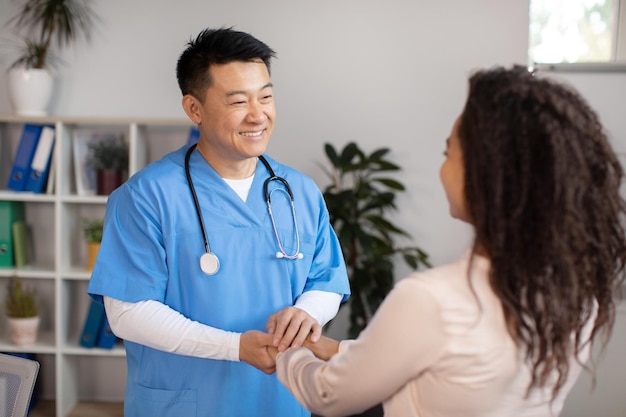 Un médecin coréen d'âge moyen joyeux se serrant la main accueille et soutient une patiente noire du millénaire