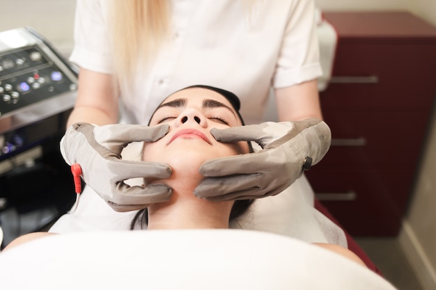 Un médecin cosmétologue fait un massage facial au patient avec des gants électriques