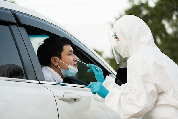 Un médecin en costume d'EPI teste le coronavirus (covid-19) à un homme en voiture par coton-tige nasal, test au volant accéléré