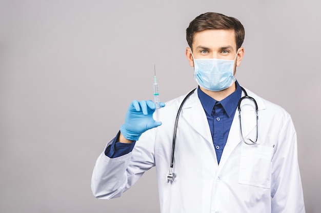 Un médecin debout levant une injection met le masque sur la bouche, accroche le stéthoscope autour du cou dans un uniforme blanc, isolé sur fond gris.