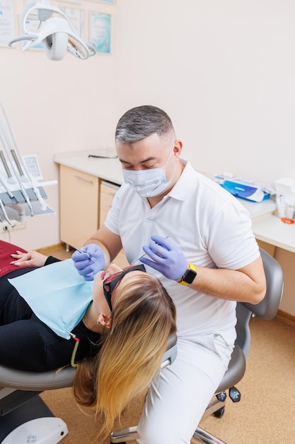 Le médecin dentiste regarde les dents du patient et tient les instruments dentaires près de la bouche L'assistant aide le médecin Ils portent des uniformes blancs avec des masques et des gants Dentiste Cabinet dentaire