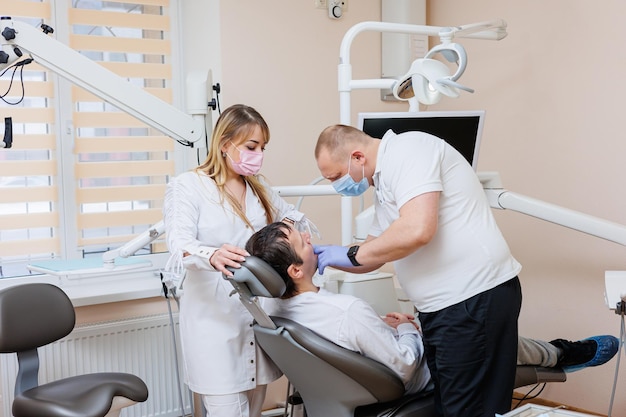 Le médecin dentiste regarde les dents des patients et tient des instruments dentaires près de la bouche L'assistant aide le médecin Ils portent des uniformes blancs avec des masques et des gants Dentiste Cabinet dentaire