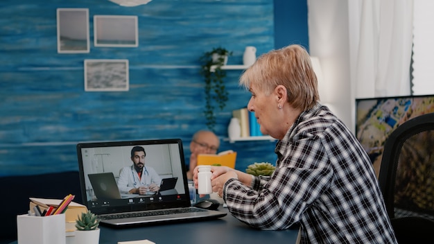 Médecin à distance utilisant une webcam prescrivant des médicaments à une femme âgée malade assise dans le salon via un stagiaire ...