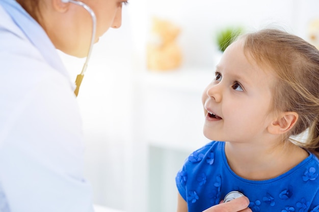 Médecin examinant un enfant au stéthoscope dans une clinique ensoleillée. Heureuse patiente souriante vêtue d'une robe bleue est à l'inspection médicale habituelle.