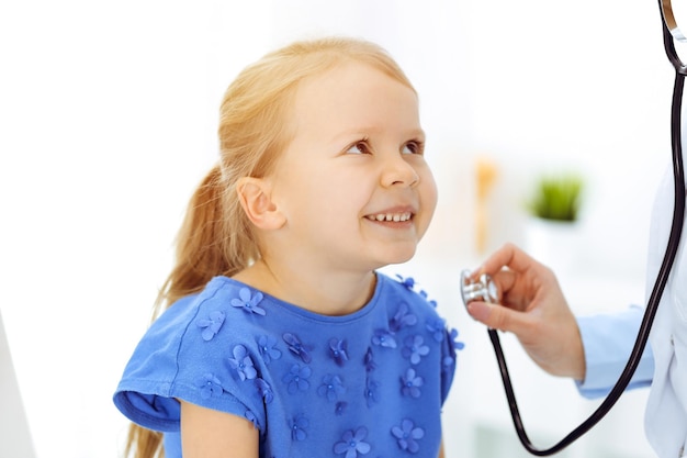 Médecin examinant un enfant au stéthoscope dans une clinique ensoleillée. Heureuse patiente souriante vêtue d'une robe bleue est à l'inspection médicale habituelle.