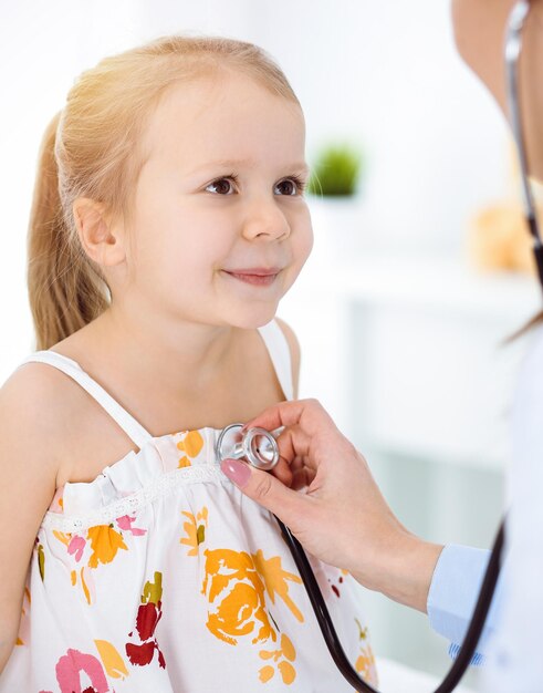 Médecin examinant un enfant au stéthoscope dans une clinique ensoleillée. Heureuse patiente souriante vêtue d'une robe de couleur vive est à l'inspection médicale habituelle.