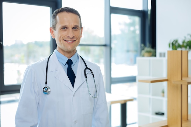 Médecin expérimenté. Homme gai sympa positif debout dans son bureau et portant un sarrau tout en travaillant comme médecin