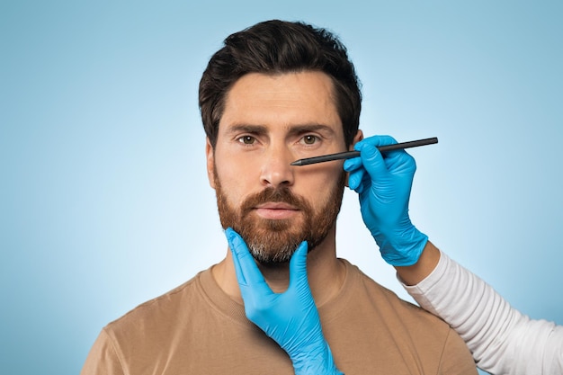 Médecin faisant des marques sur le visage masculin bel homme caucasien en consultation chez le chirurgien debout sur fond bleu