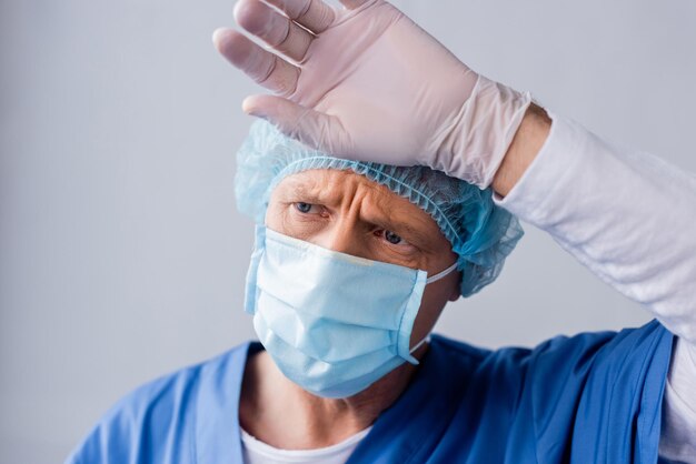 Médecin fatigué et mature en masque médical bleu et casquette touchant le front sur gris