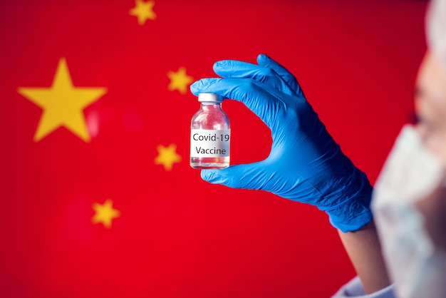 Un médecin en gants médicaux bleus et masque tenant une ampoule avec un vaccin devant le drapeau de la Chine. Photo en gros plan. Coronavirus, épidémie et concept de médecine.