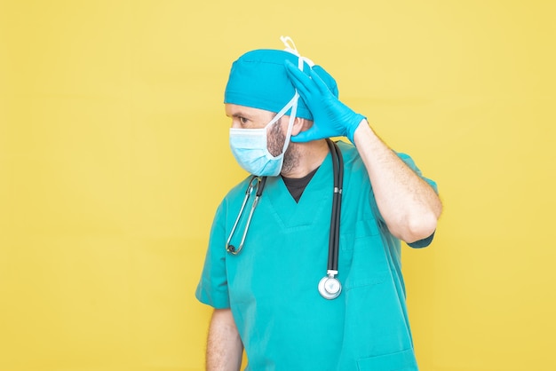 Médecin habillé en chirurgien en vert avec stéthoscope et masque sur fond jaune avec une expression inquiète.