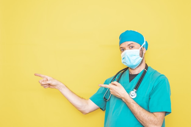 Médecin habillé en chirurgien en vert avec stéthoscope et masque sur fond jaune avec un geste pointant vers une direction.