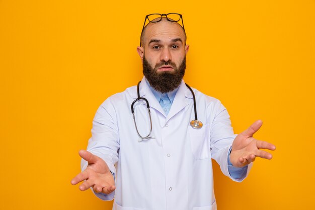 Médecin homme barbu en blouse blanche avec stéthoscope autour du cou avec des lunettes sur la tête regardant la caméra confus levant les bras de mécontentement debout sur fond orange