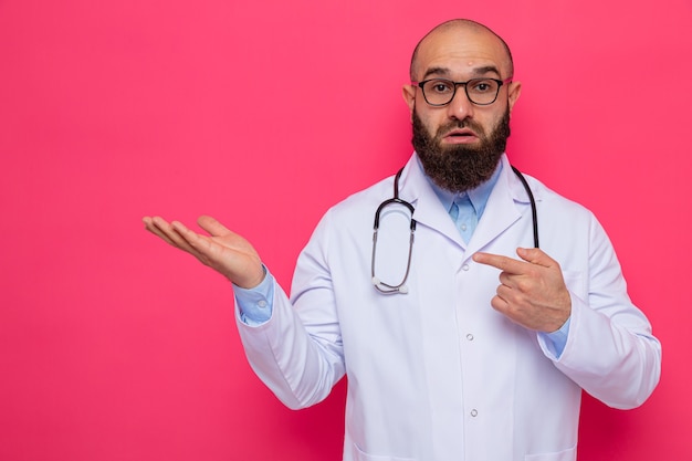 Médecin homme barbu en blouse blanche avec stéthoscope autour du cou portant des lunettes confus pointant avec l'index sur le côté présentant quelque chose avec le bras de sa main