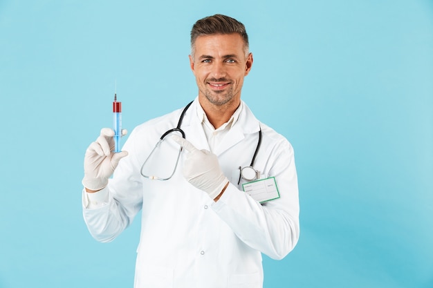 Médecin homme confiant en uniforme debout isolé sur mur bleu, montrant une seringue avec un liquide rouge