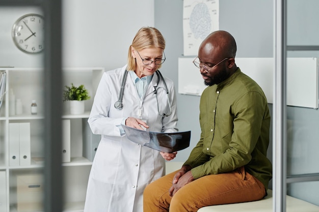 Médecin mature confiant en blouse blanche montrant une image radiographique d'un jeune homme noir