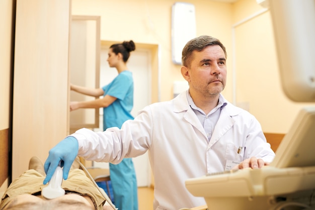 Photo médecin mature sérieux en blouse de laboratoire faisant une échographie lors de l'examen de l'abdomen du patient