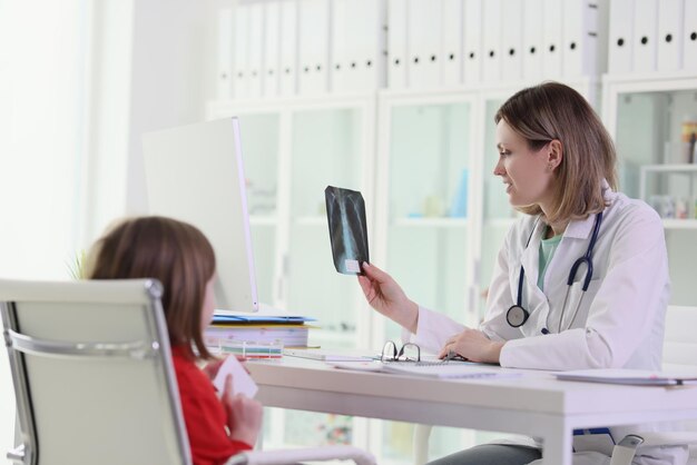 Un médecin montre une image radiographique des poumons à une petite fille assise à table femme en uniforme médical