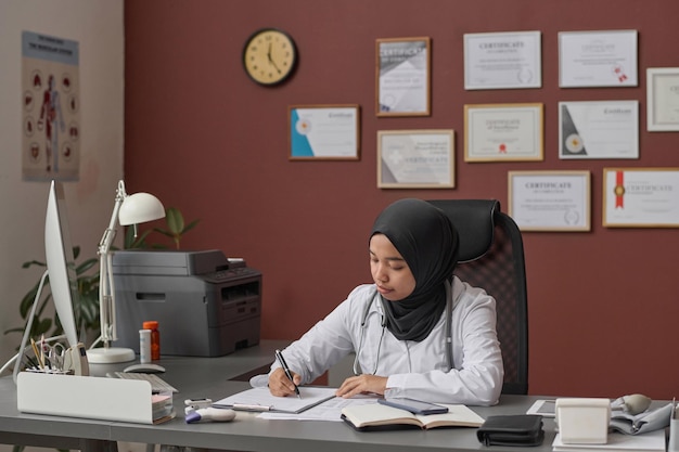 Un médecin musulman écrivant dans un dossier médical assis au bureau de l'hôpital