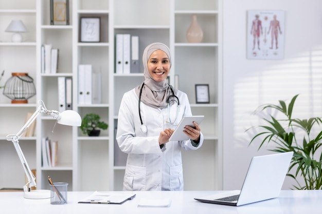 Une médecin musulmane souriante en hijab et manteau blanc utilisant une tablette dans un bureau médical lumineux