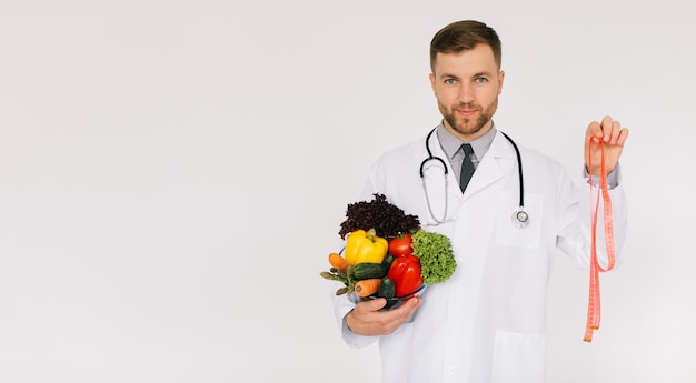 Le médecin nutritionniste masculin avec stéthoscope tenant des légumes frais et une règle sur fond blanc