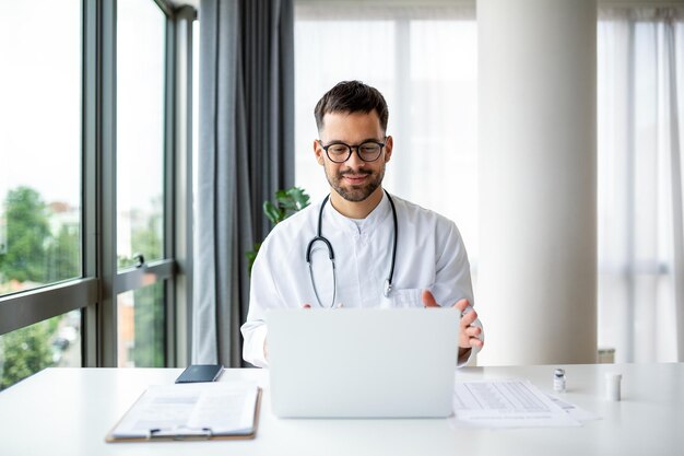 Médecin avec ordinateur portable effectue un appel vidéo en ligne et parle au patient Médecin de sexe masculin travaillant à l'intérieur du bureau de bureau sur fond