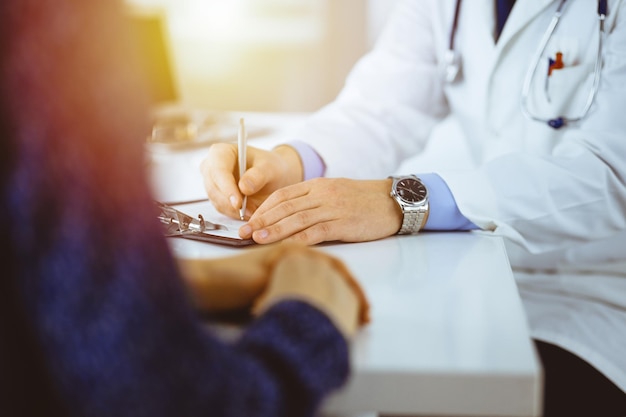 Un médecin parle à son patient, assis ensemble au bureau dans le cabinet ensoleillé d'un hôpital. Médecin utilisant le presse-papiers pour remplir les dossiers d'historique des médicaments. Service médical parfait