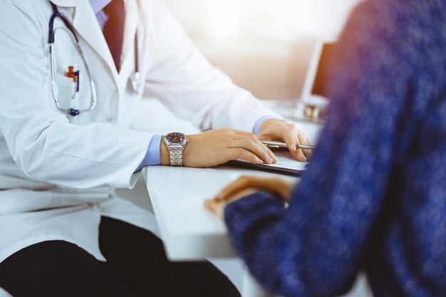 Un médecin parle à son patient, assis ensemble au bureau dans le cabinet ensoleillé d'un hôpital. Médecin utilisant le presse-papiers pour remplir les dossiers d'historique des médicaments. Service médical parfait