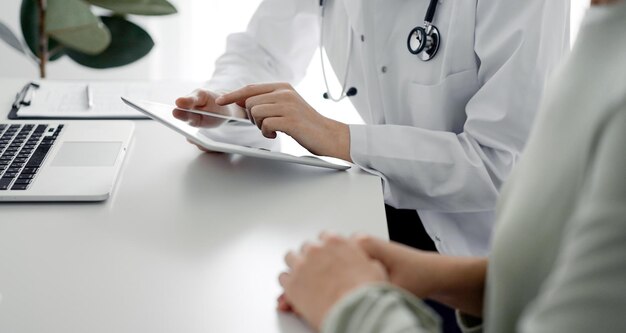 Médecin et patient assis au bureau du bureau de la clinique. L'accent est mis sur les mains de la femme médecin à l'aide d'un ordinateur tablette, gros plan. Notion de médecine.