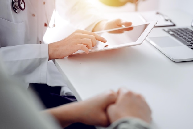 Médecin et patient assis au bureau du bureau de la clinique. L'accent est mis sur les mains de la femme médecin à l'aide d'un ordinateur tablette, gros plan. Notion de médecine.