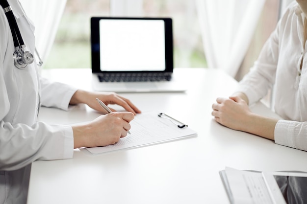 Médecin et patient assis l'un près de l'autre au bureau blanc de la clinique. Une femme médecin écoute en remplissant un formulaire d'enregistrement. Notion de médecine.