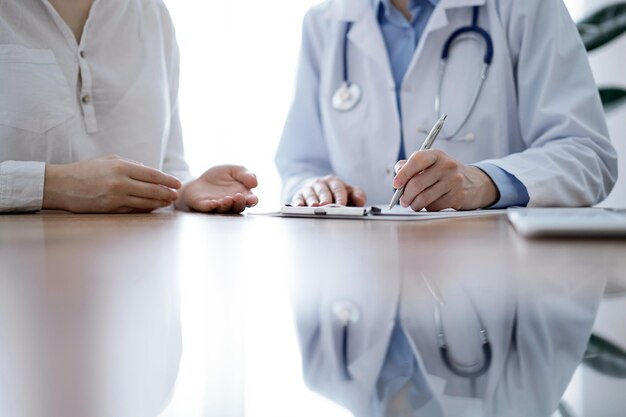 Médecin et patient assis l'un près de l'autre au bureau en bois de la clinique. Une femme médecin remplit un formulaire d'enregistrement. Notion de médecine