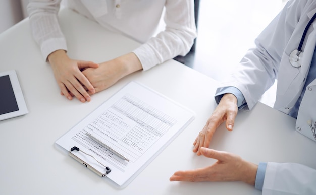 Médecin et patient discutant de questions de santé actuelles tout en étant assis l'un à côté de l'autre et en utilisant le presse-papiers à la table de la clinique, juste les mains en gros plan. Notion de médecine.