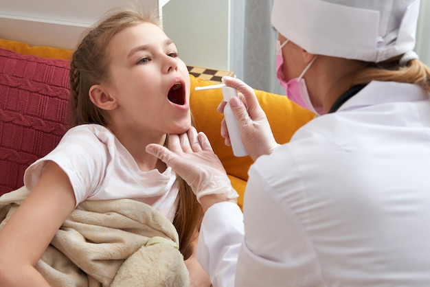 Médecin pédiatre à l'aide d'un spray médical pour jeune fille à la maison, mal de gorge