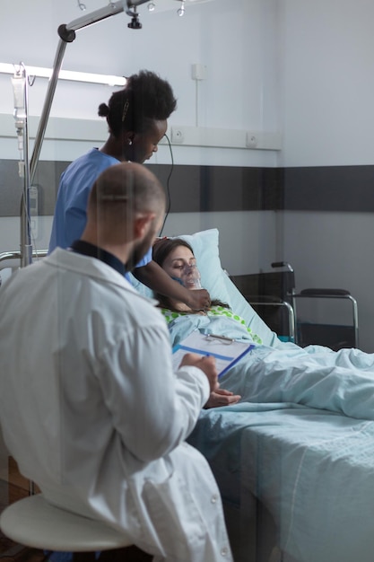 Médecin pendant les rondes du matin vérifiant le presse-papiers avec les dossiers des patients pendant que l'infirmière met un masque à oxygène sur une femme inconsciente. Soignant assistant une femme en convalescence dans une salle d'hôpital moderne.