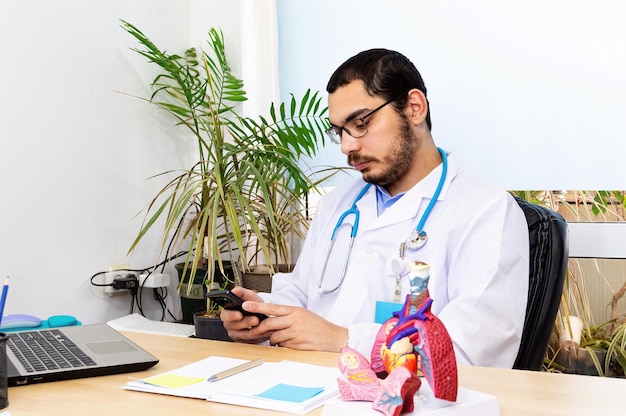 Médecin pneumologue vérifiant un téléphone intelligent dans un bureau moderne et un modèle anatomique du poumon sur le bureau