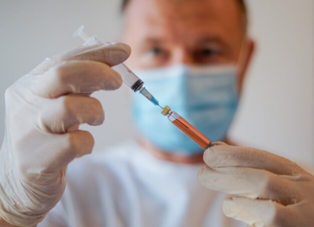 Médecin portant des gants de protection et un masque facial gardant dans sa main une injection avec un vaccin, un antidote et un traitement pour la pandémie de virus Covid-19
