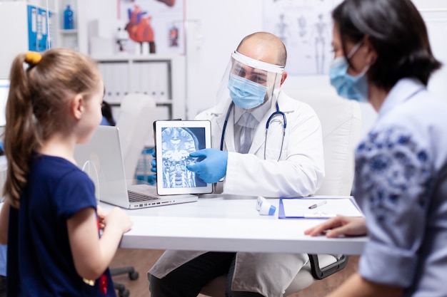 Médecin portant un masque facial pointant sur une radiographie sur une tablette pendant la consultation d'un enfant