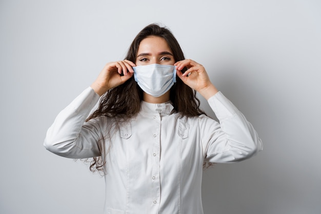 Médecin portant un masque médical sur fond blanc. Jeune femme séduisante en robe médicale. Tendances de la mise en quarantaine du coronavirus covid-19.