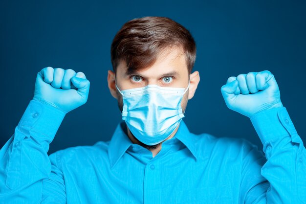 Un médecin portant un masque médical et des gants, en uniforme bleu, fait des gestes avec ses mains.