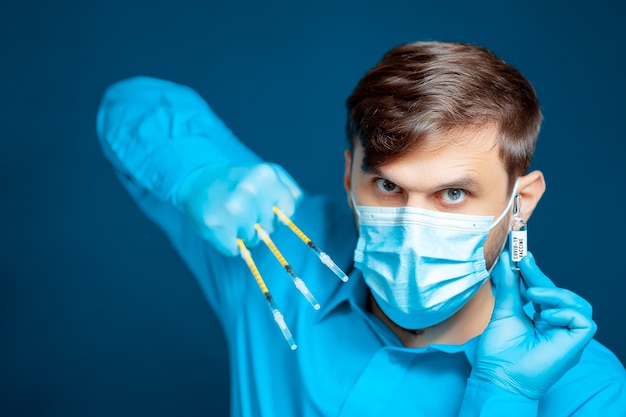 Un médecin portant un masque médical et des gants, en uniforme bleu, pose avec de fines seringues entre ses doigts devant la caméra