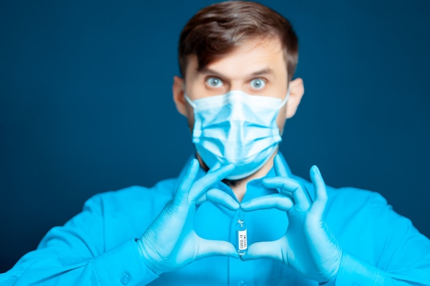 Un médecin portant un masque médical et des gants, en uniforme bleu, tient des ampoules contenant un vaccin contre le coronavirus devant lui avec ses doigts.