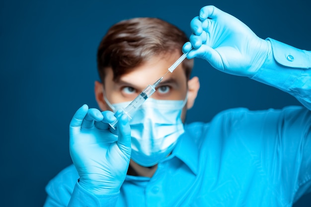 Un médecin portant un masque médical et des gants vêtus d'un uniforme bleu enlève le capuchon de l'aiguille de la seringue