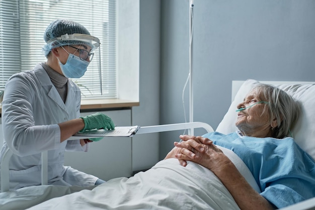 Médecin portant un masque de protection et des gants prenant des notes sur la carte médicale tout en examinant une femme âgée allongée sur son lit dans le service