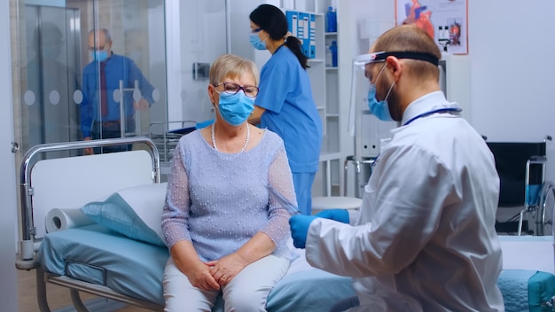 Médecin portant une visière vérifiant la radiographie des poumons d'une vieille femme âgée à la retraite dans une clinique hospitalière privée moderne. Établissement de santé pendant la pandémie mondiale de COVID-19, portant un équipement de protection. Médical