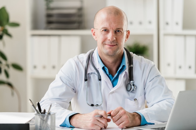 Médecin professionnel chauve avec stylo assis par bureau dans un cabinet médical ou à l'hôpital et effectuant son travail