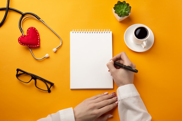 Médecin professionnel écrivant des dossiers médicaux dans un cahier avec stéthoscope, tasse à café, seringue et coeur