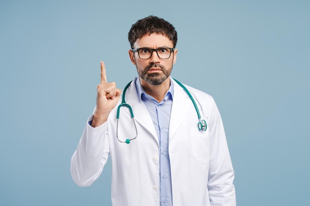Un médecin professionnel sérieux en colère portant un manteau blanc et des lunettes pointant le doigt vers le haut