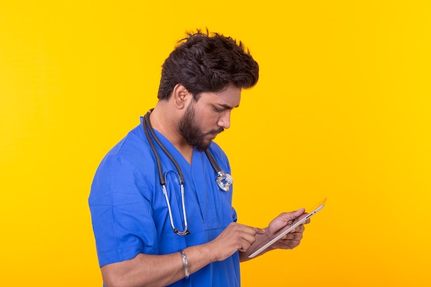 Médecin de race mixte mignon dans une robe bleue avec un stéthoscope en regardant une tablette basée sur un mur jaune. Consultation en ligne de concept. Copyspace