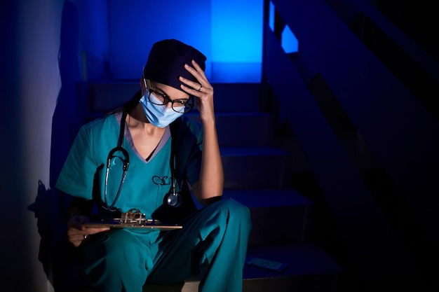 Une médecin réfléchie en masque de protection et en uniforme assise sur l'escalier avec un bloc-notes tout en lisant des documents et en touchant la tête